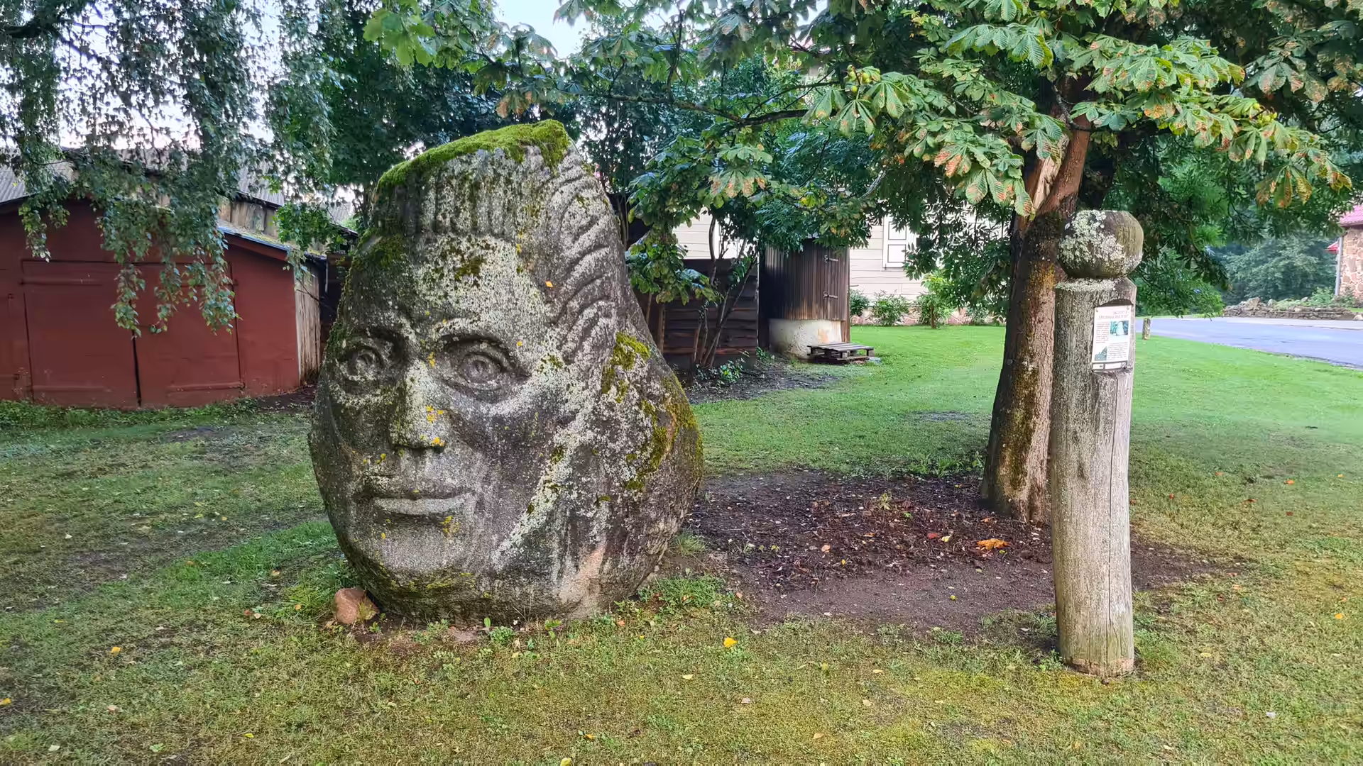 A large stone sculpture carved with a human face standing beneath trees in a peaceful village in Lithuania.