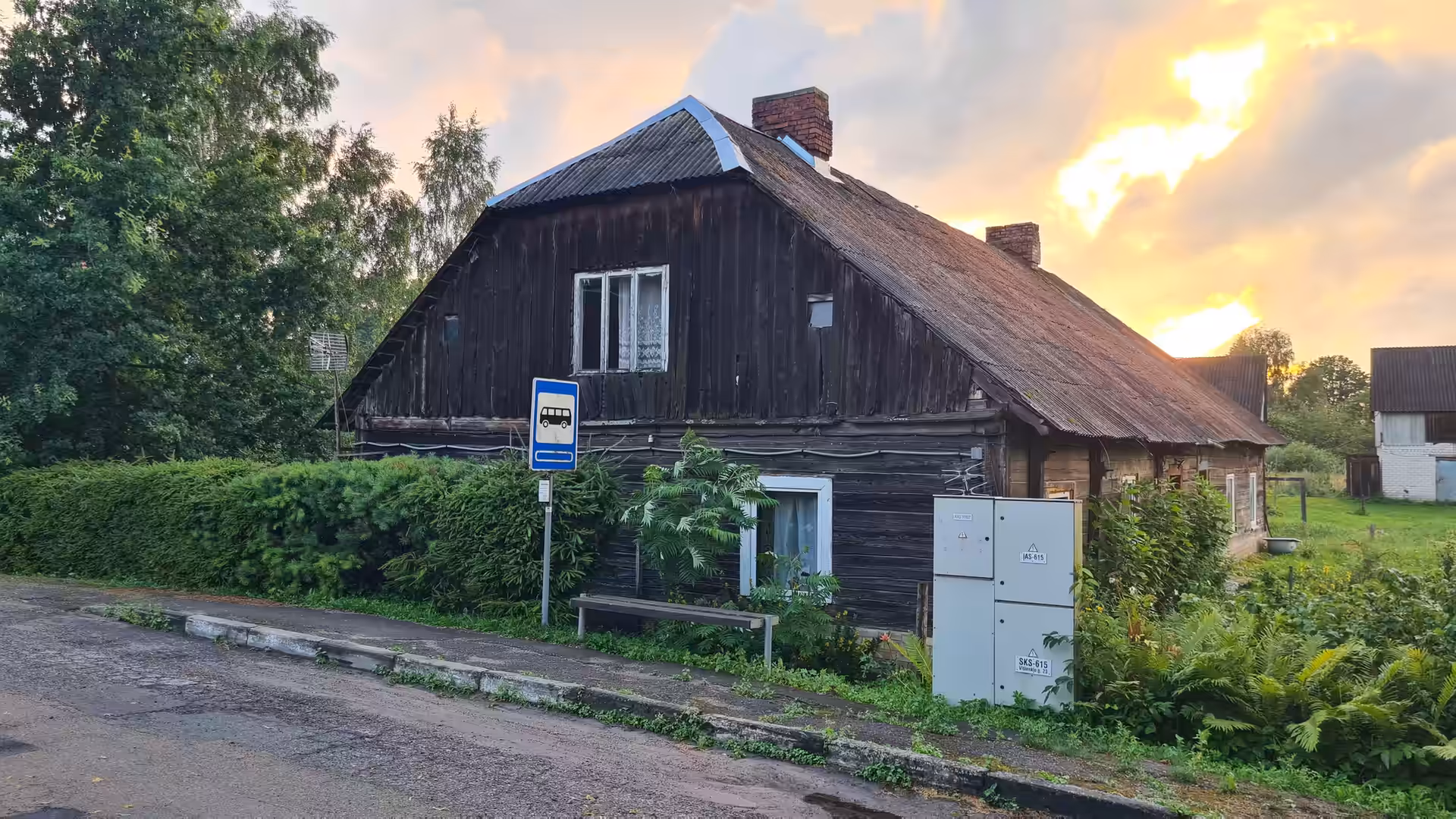 An old wooden farmhouse beside a quiet village road in Lithuania, illuminated by a colorful sunset after rainfall.