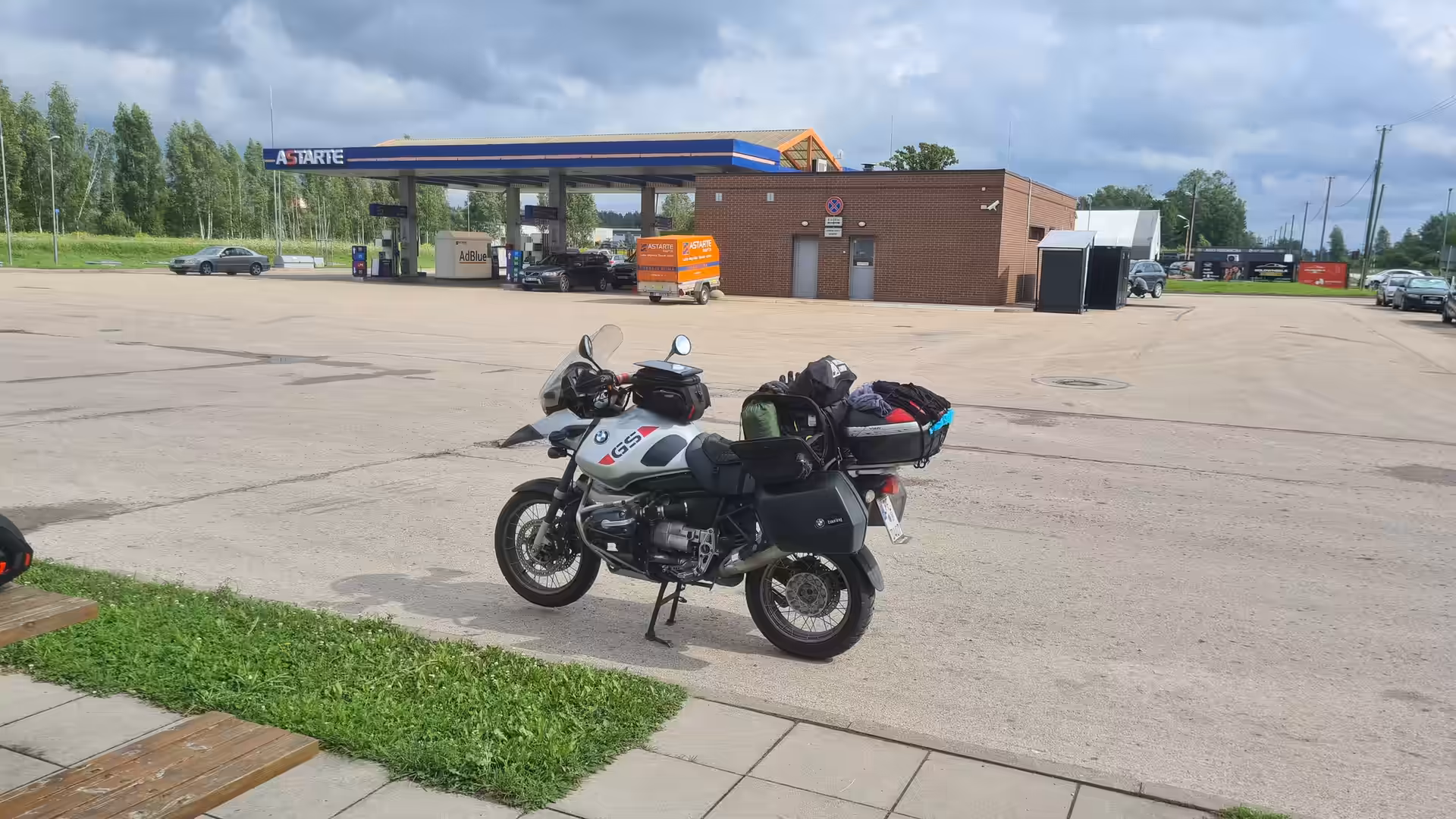A fully loaded BMW GS adventure motorcycle parked at a fuel station in Latvia beneath dramatic storm clouds.