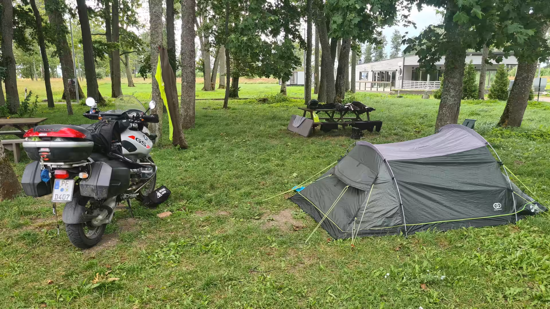 A fully loaded BMW GS adventure motorcycle parked beside a small backpacking tent at a shaded campsite in Estonia.