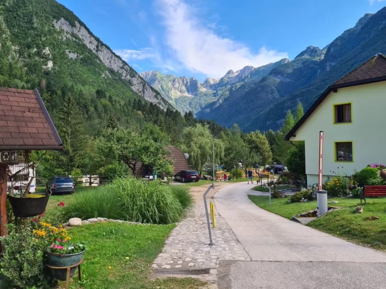 A stone-paved path curves through a green alpine village surrounded by high mountains.