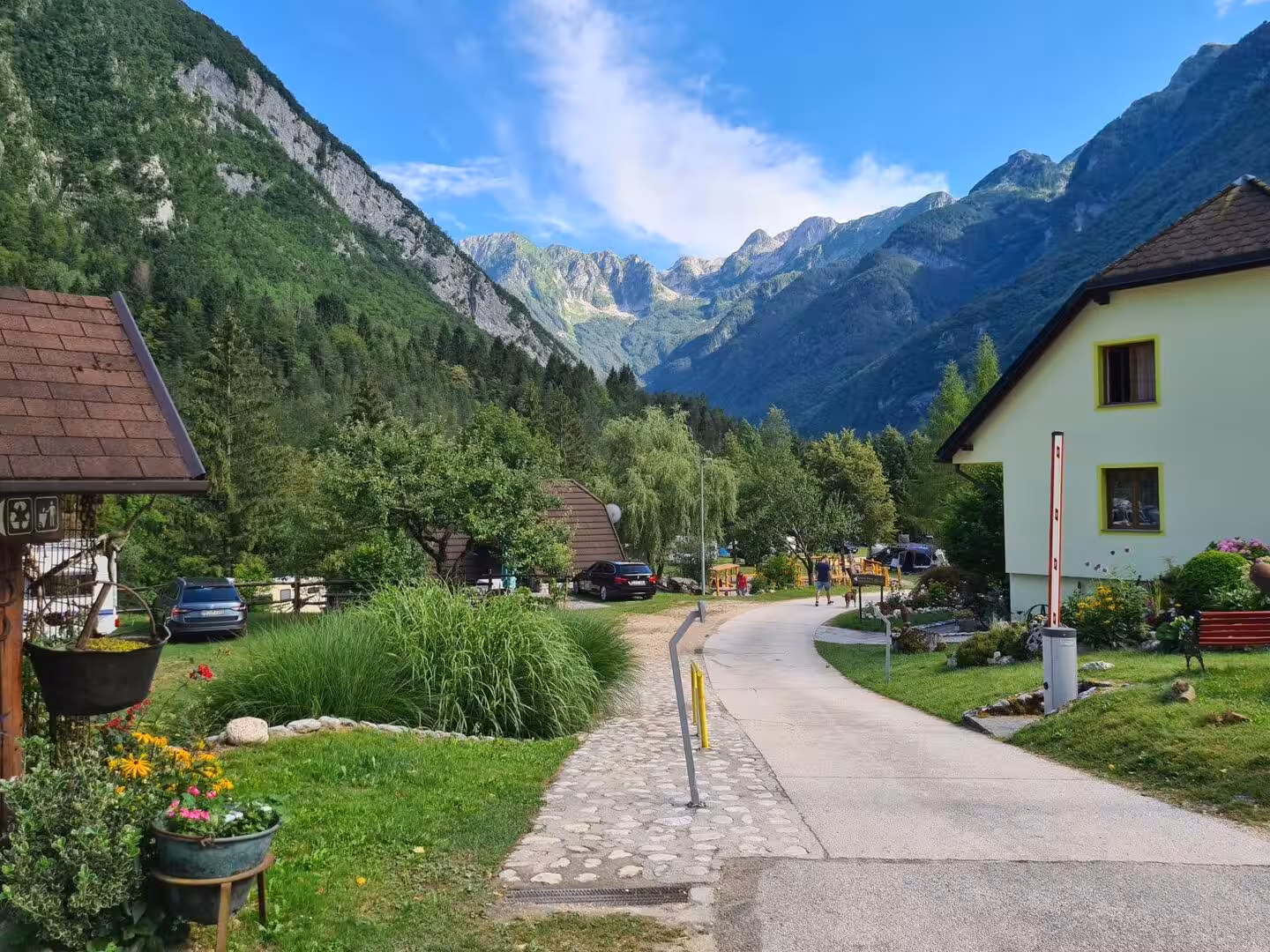 A stone-paved path curves through a green alpine village surrounded by high mountains.