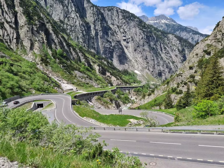 A winding mountain road with hairpin bends climbs through a green gorge beneath a snow-capped peak.