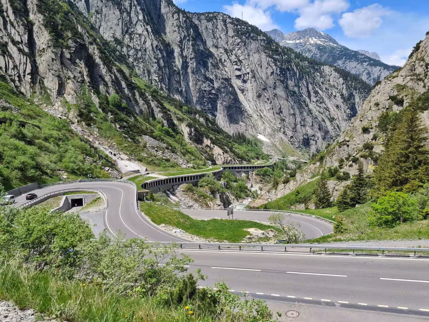 A winding mountain road with hairpin bends climbs through a green gorge beneath a snow-capped peak.