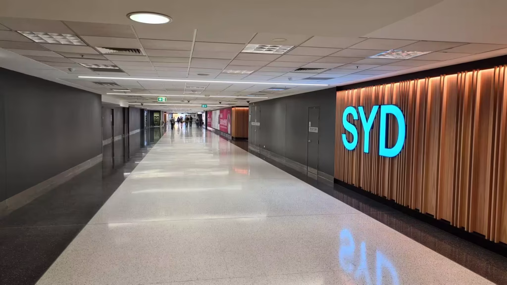 Blue SYD sign glows on a timber slat wall in a bright arrivals corridor.