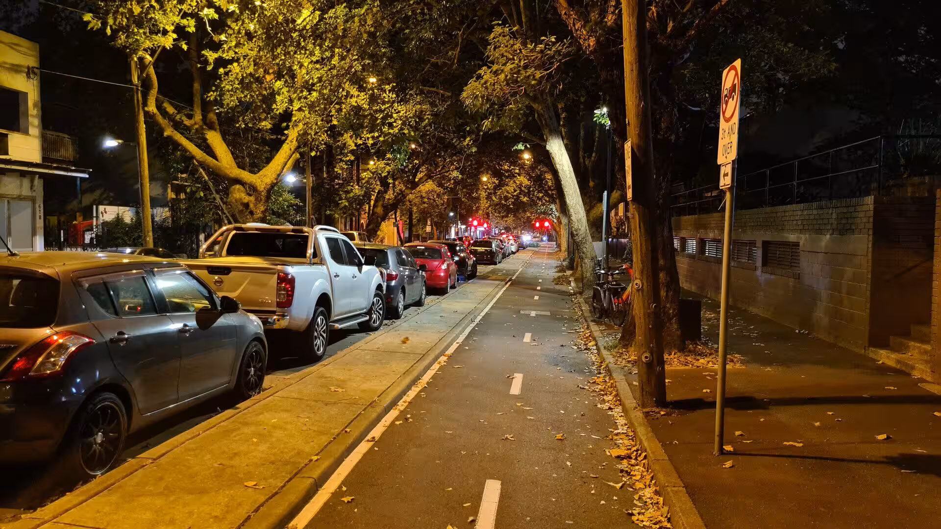 A warm-lit residential street at night, with cars parked under leafy trees.
