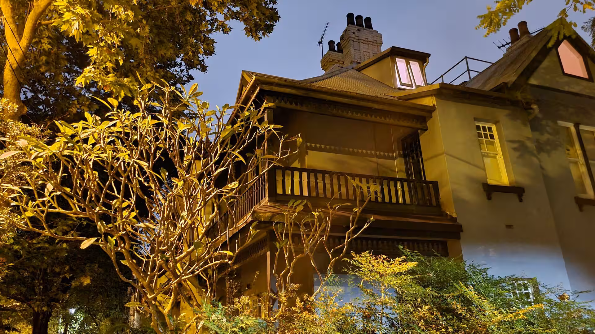 A classic two-storey home with a wooden balcony and multiple chimneys glows at dusk.