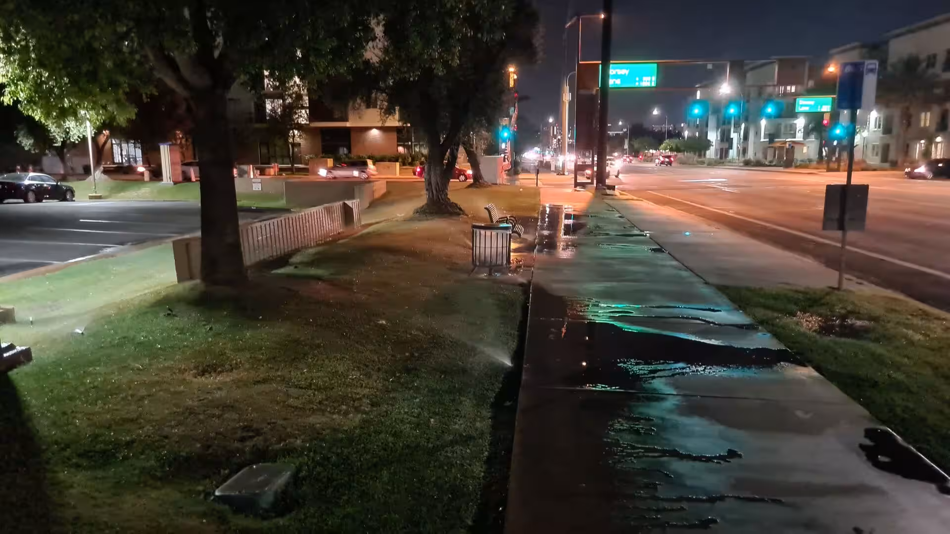 A rain-wet sidewalk reflects the glow of a busy street at night.