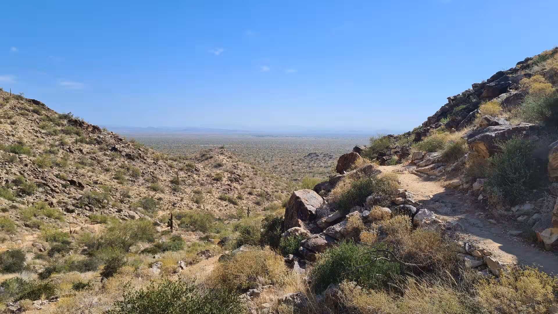 A rocky desert trail opens onto a sweeping flat valley reaching to the horizon.