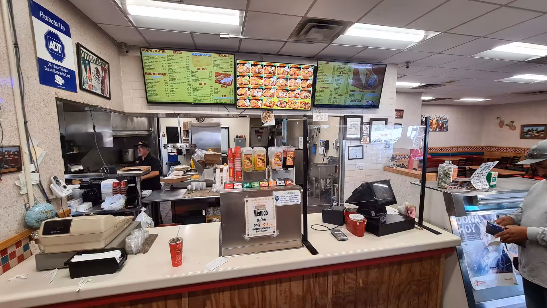 A Mexican food counter with taco menu boards and a cook in back