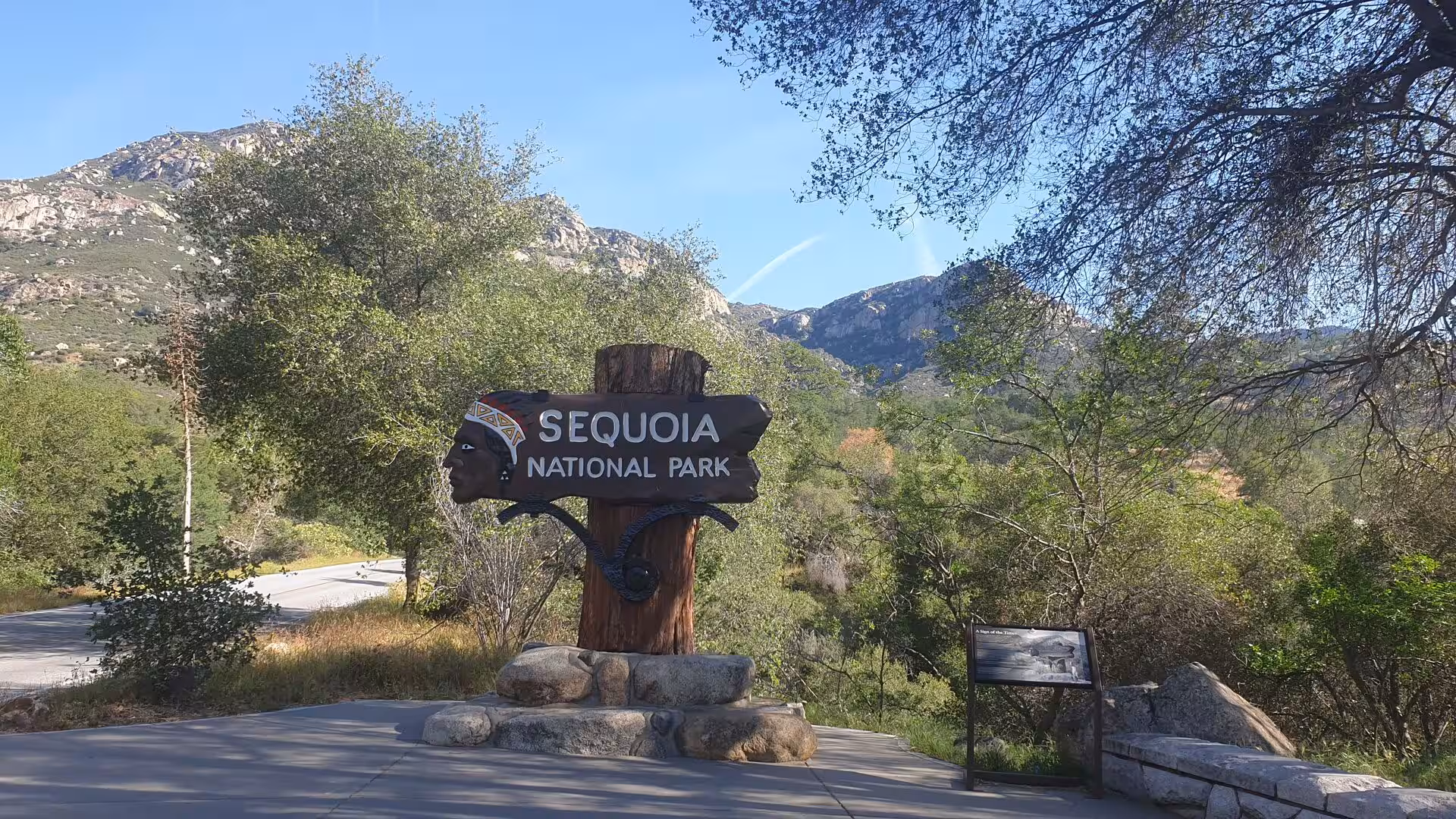 A carved wooden Sequoia National Park sign stands at the roadside surrounded by green trees and rocky foothills.