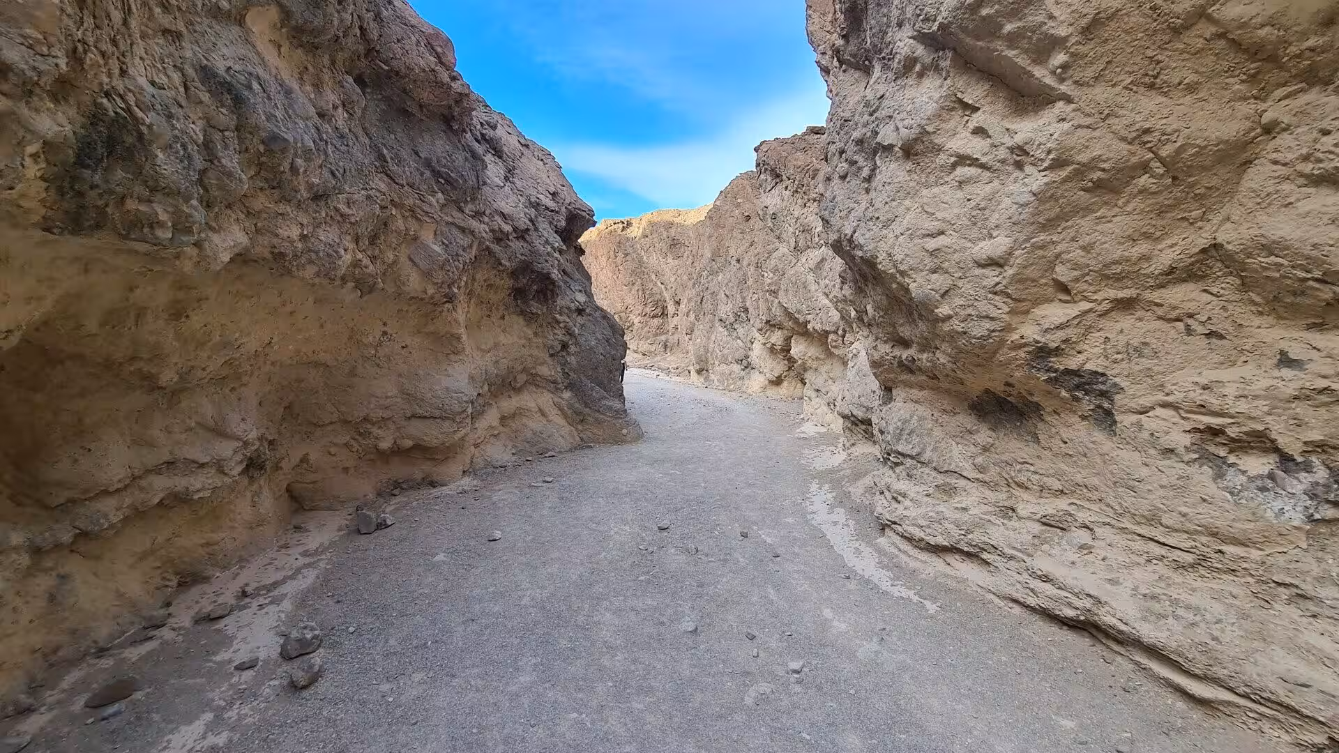 A dry sandy floor cuts between towering tan rock walls beneath a strip of blue sky