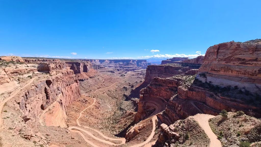 A narrow dirt road switchbacks down sheer red sandstone walls into a deep desert gorge.