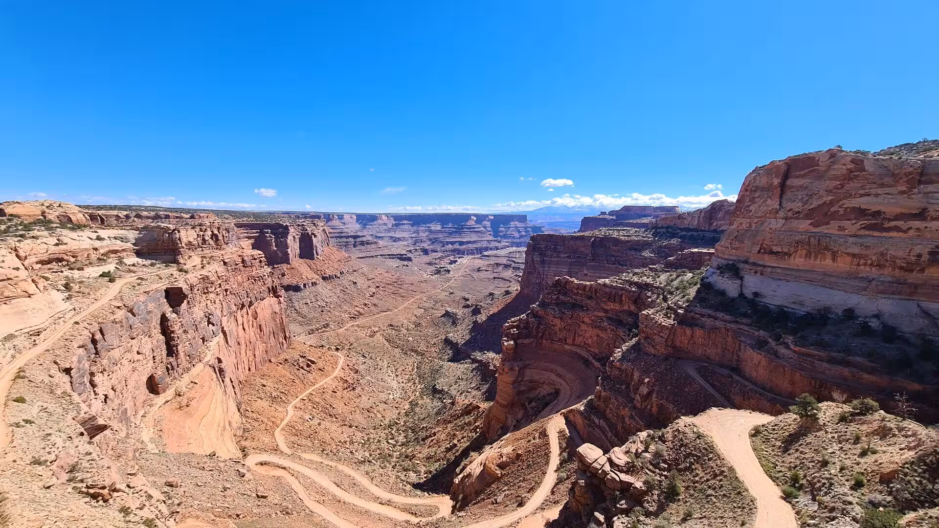 A narrow dirt road switchbacks down sheer red sandstone walls into a deep desert gorge.