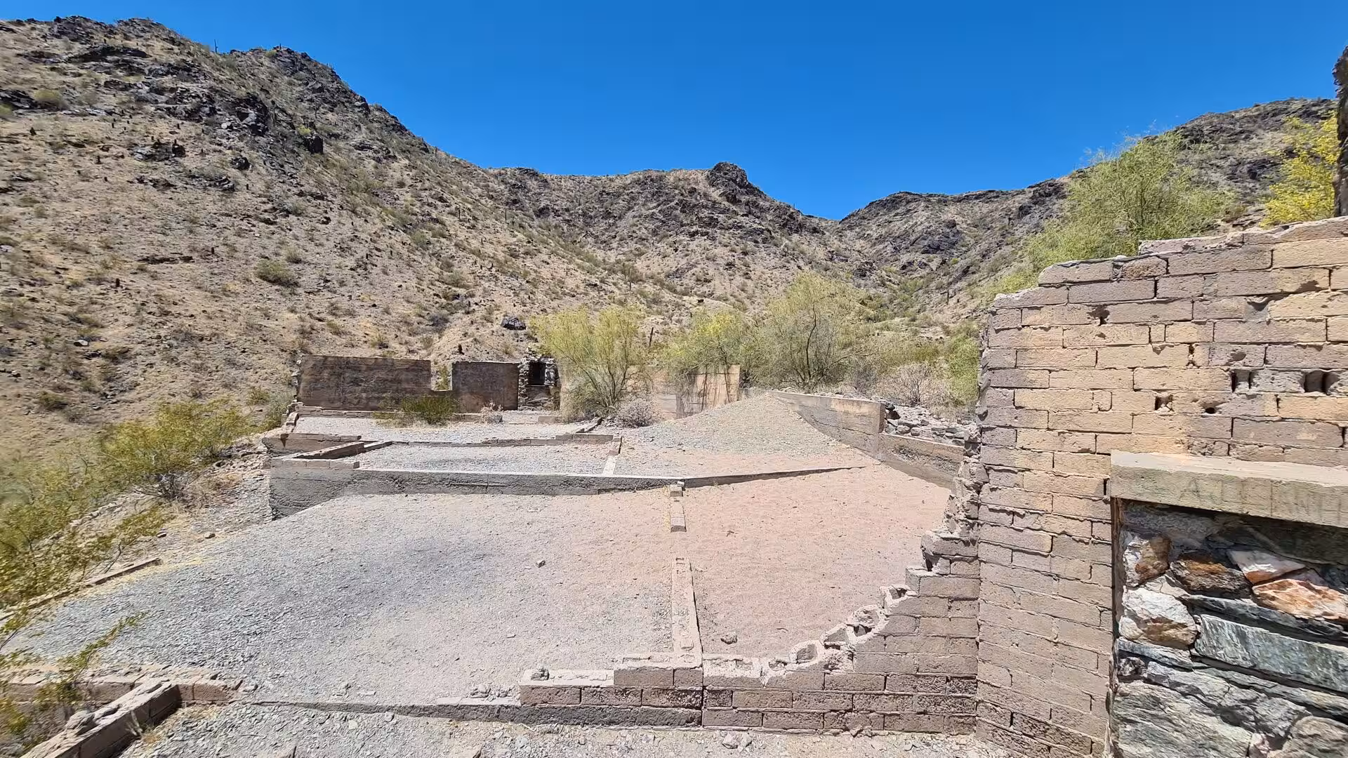 Low stone walls sit among green trees in a dry desert valley.