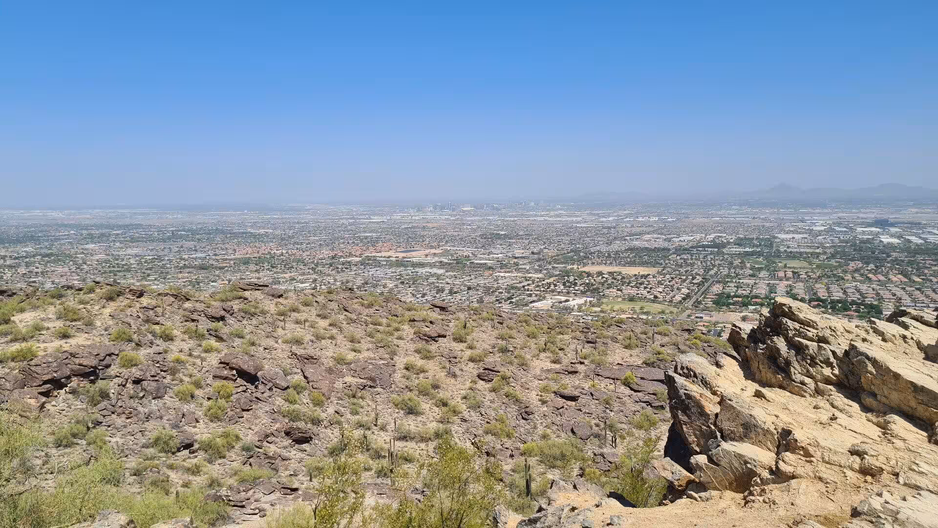 A sprawling city spreads out beneath a hazy blue sky from a rocky desert summit.