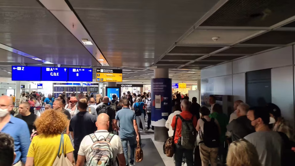 Passengers with backpacks stream through a busy airport hall beneath blue directional signs