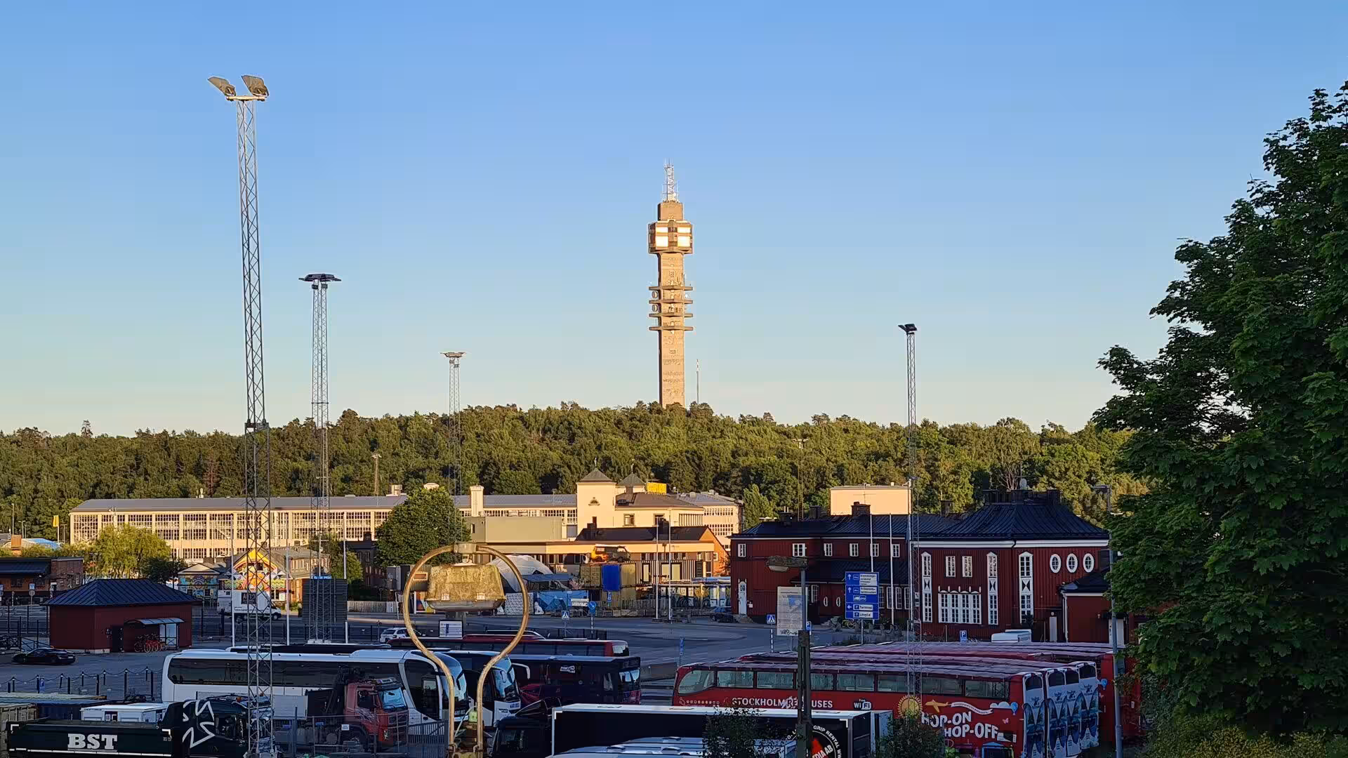 Red hop-on hop-off buses wait in a car park beneath a tower over the trees.