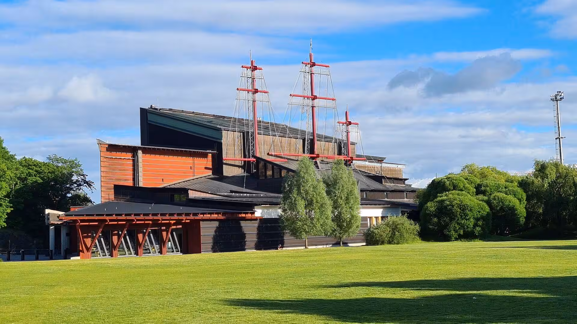 A contemporary building is framed by three towering red ship masts.
