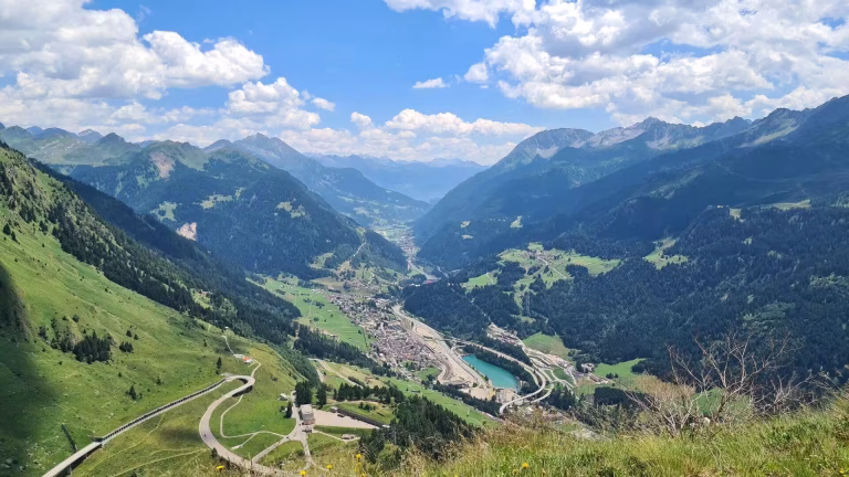A winding road crosses a green alpine valley with a turquoise lake below.