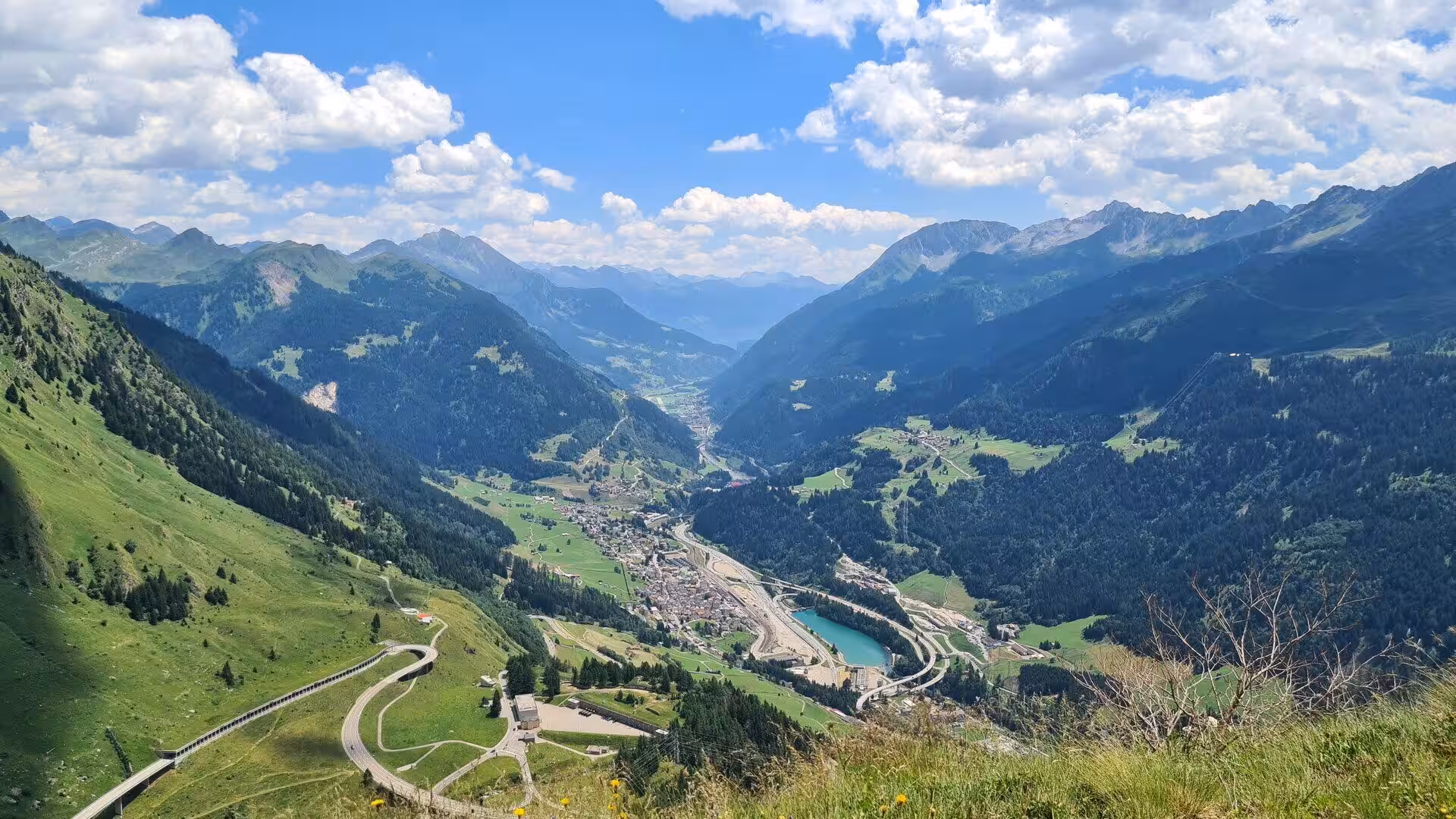 A winding road crosses a green alpine valley with a turquoise lake below.