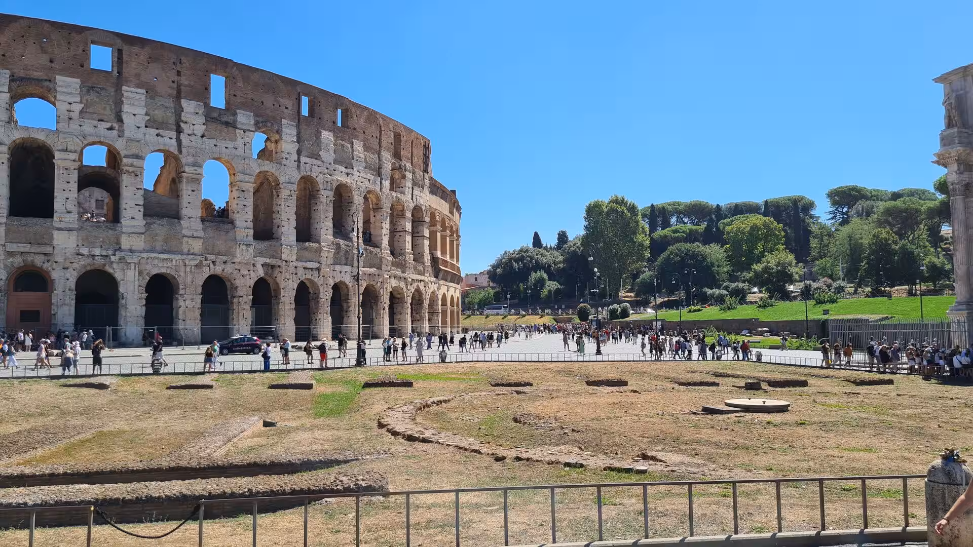 The huge arched ruins of the Colosseum tower over crowds strolling across an open plaza in bright sun.