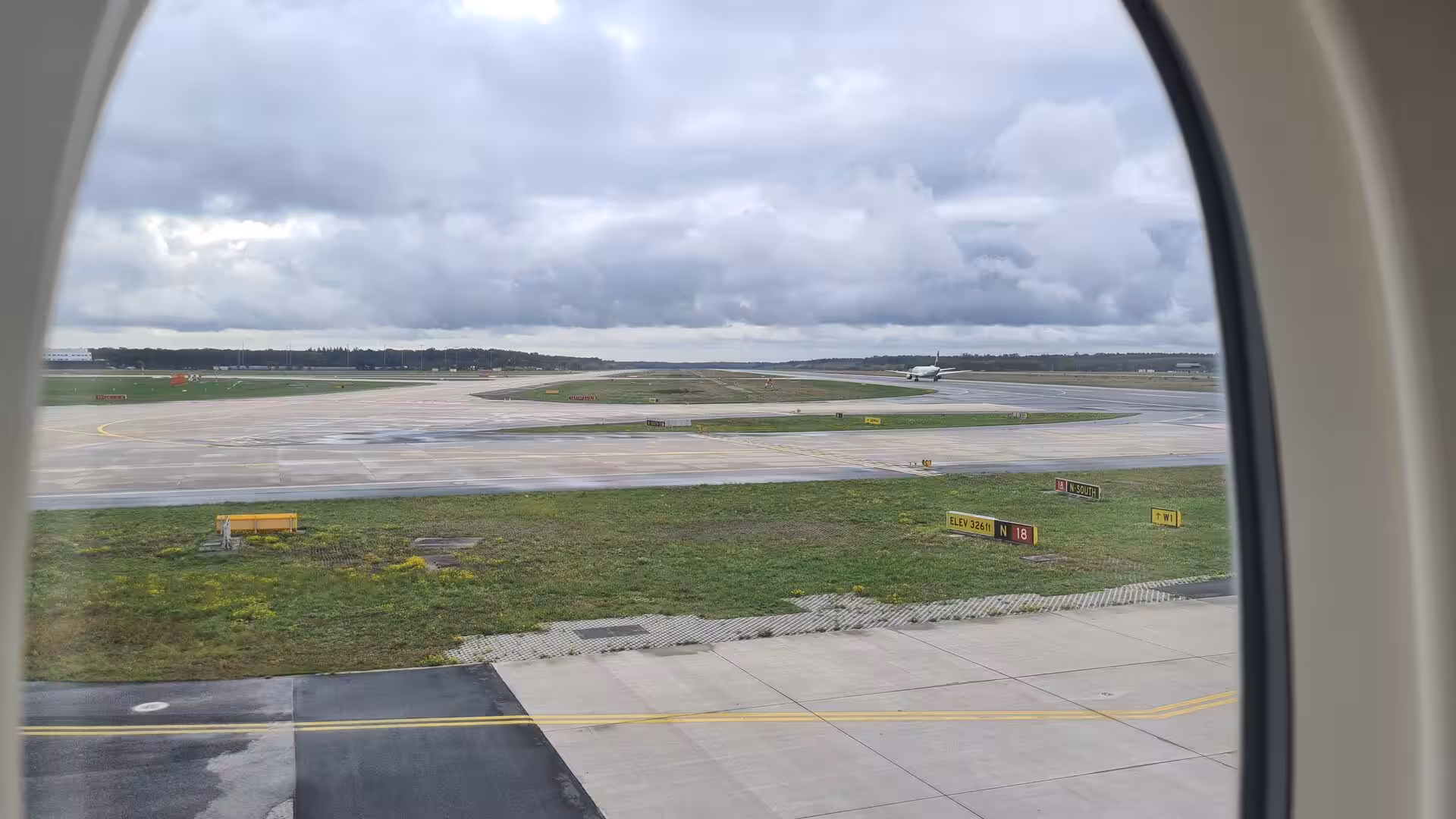 Tarmac and green grass with runway signs stretch below, framed by the rounded cabin window.