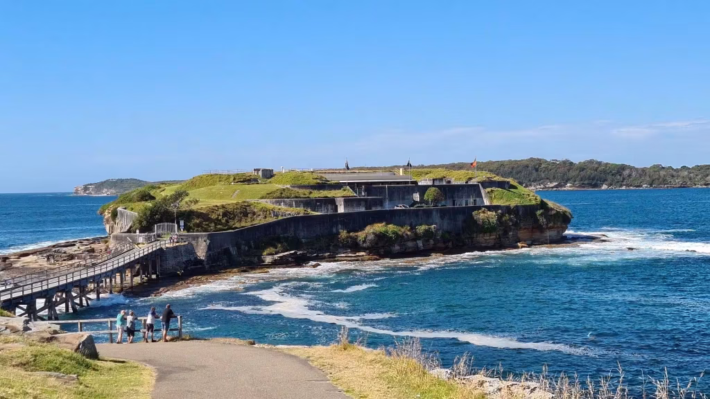 A green-topped sea headland crowned by a weathered concrete fort juts into the blue water.