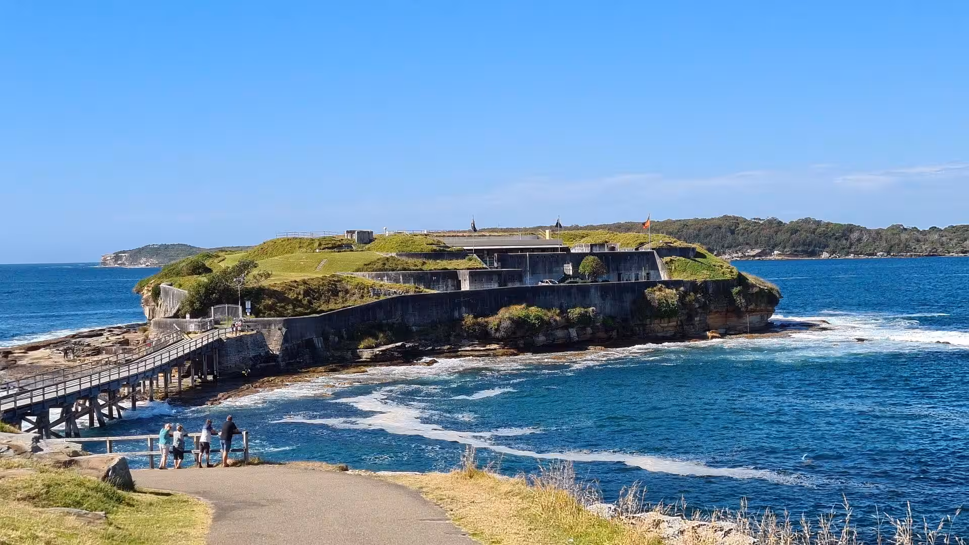 A green-topped sea headland crowned by a weathered concrete fort juts into the blue water.