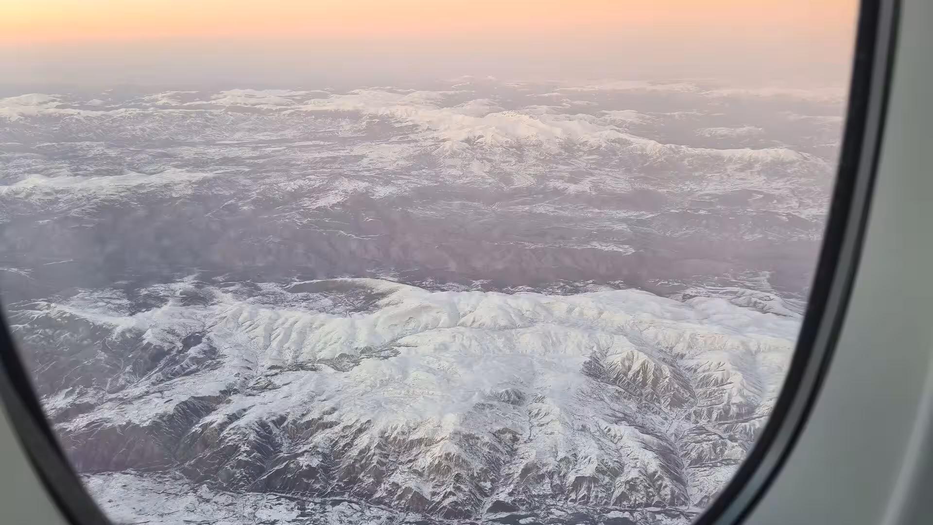 Rugged snow-covered mountains stretch to a warm pink horizon seen through a plane window.
