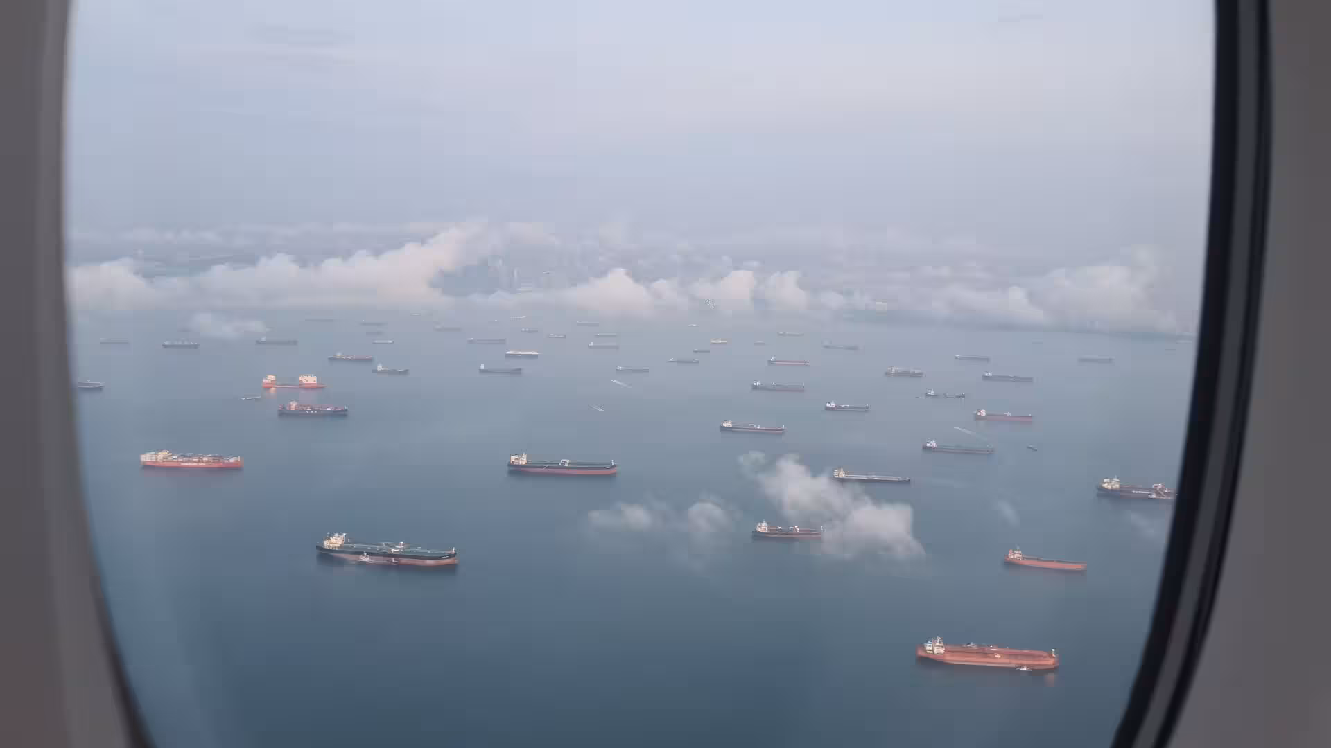 Dozens of tankers and cargo ships rest at anchor across a hazy bay near a distant coastline.