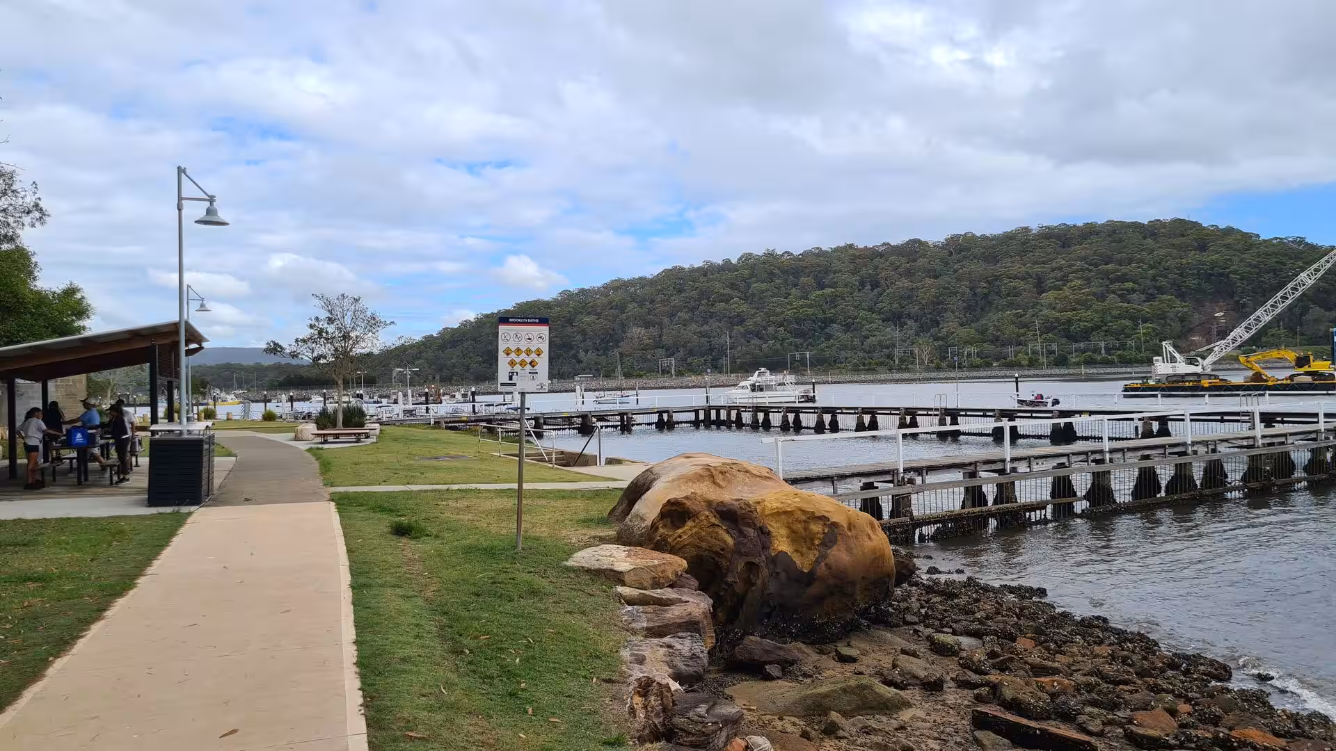 A waterfront path winds past a picnic shelter, a large boulder and a boat-filled pier.