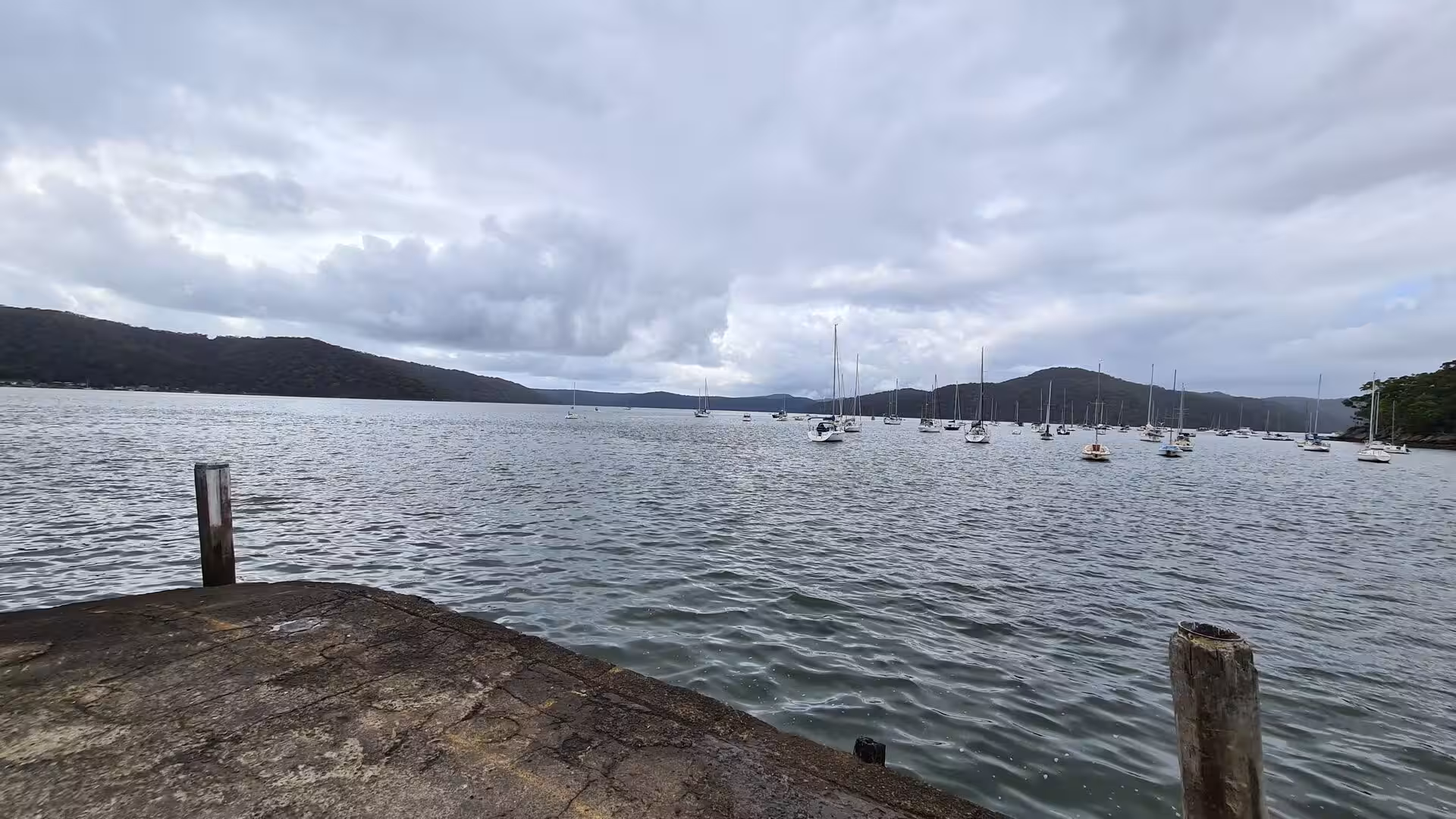 Many sailboats with tall masts rest in calm water, seen from a low stone wharf.