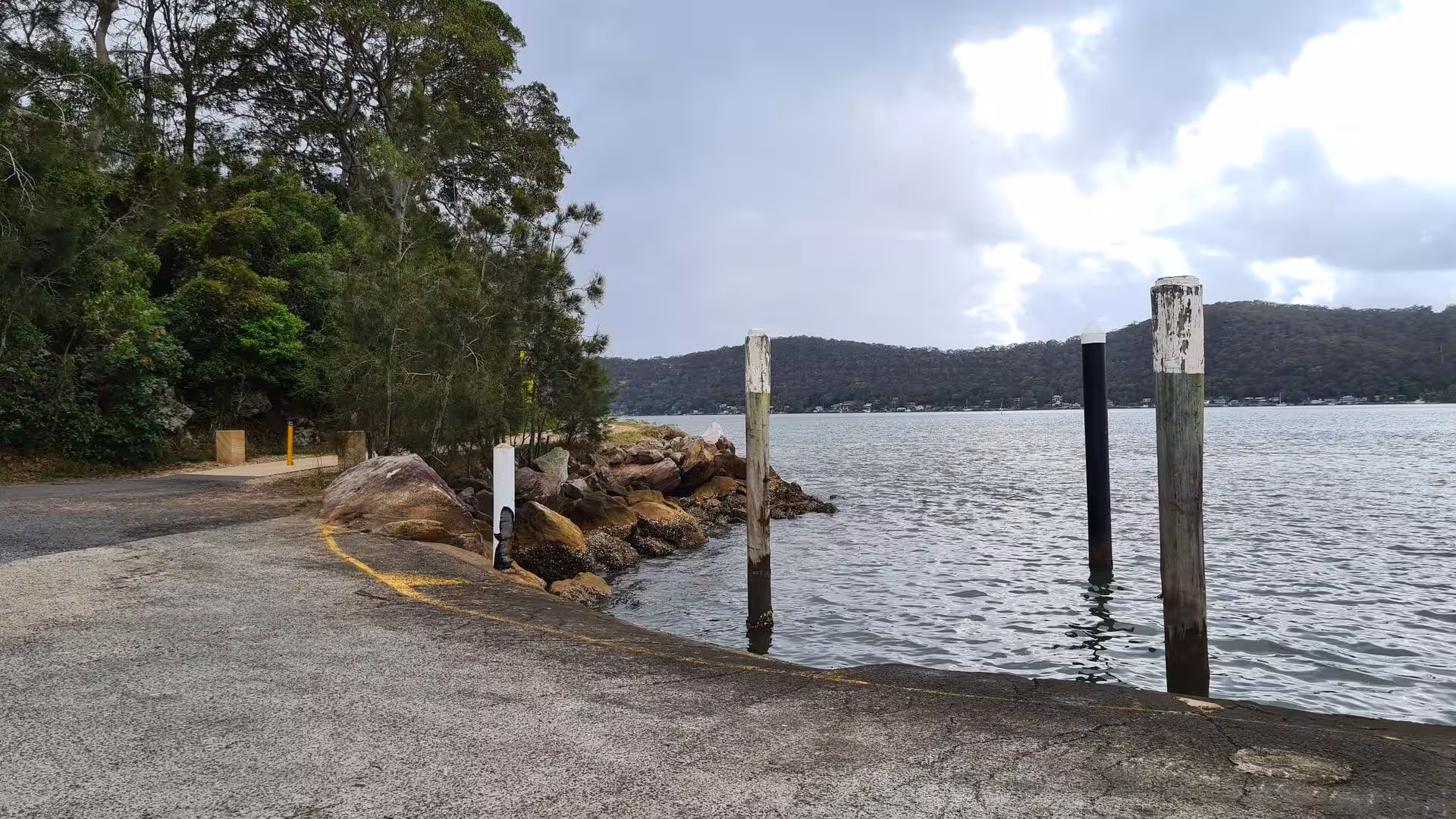 Wooden mooring poles flank a concrete boat ramp that slips into still water.