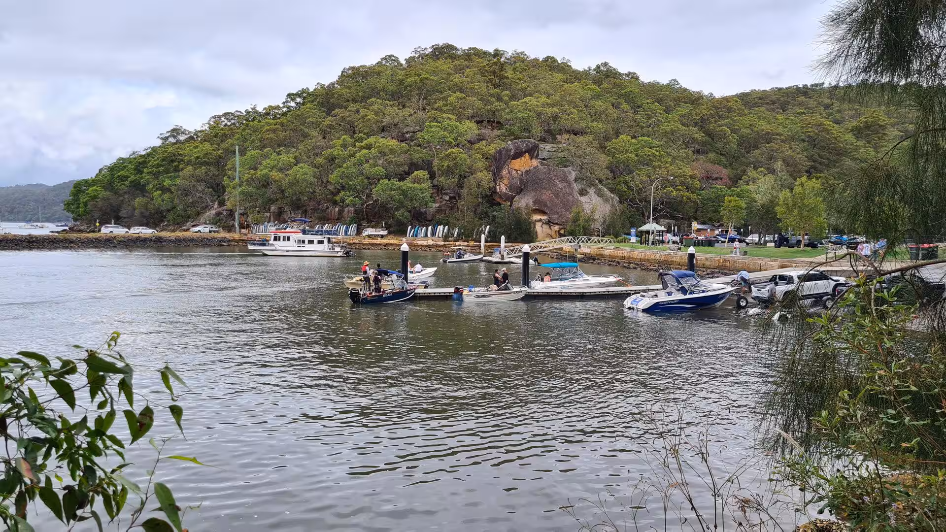 Boats tie up at a pontoon below a forested hill marked by a large orange rock overhang.