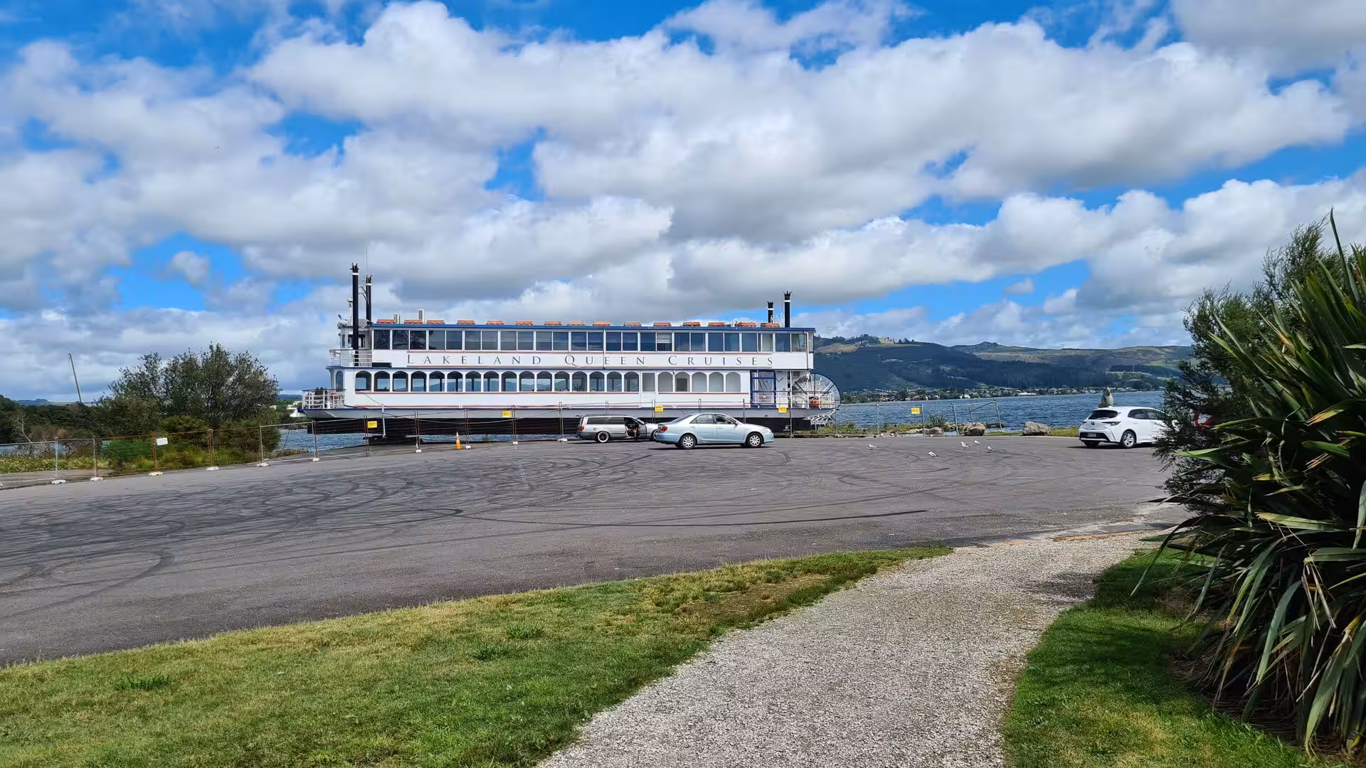 A white two-deck paddle steamer marked 'Lakeland Queen Cruises' sits moored at a lakeside pier beside parked cars.
