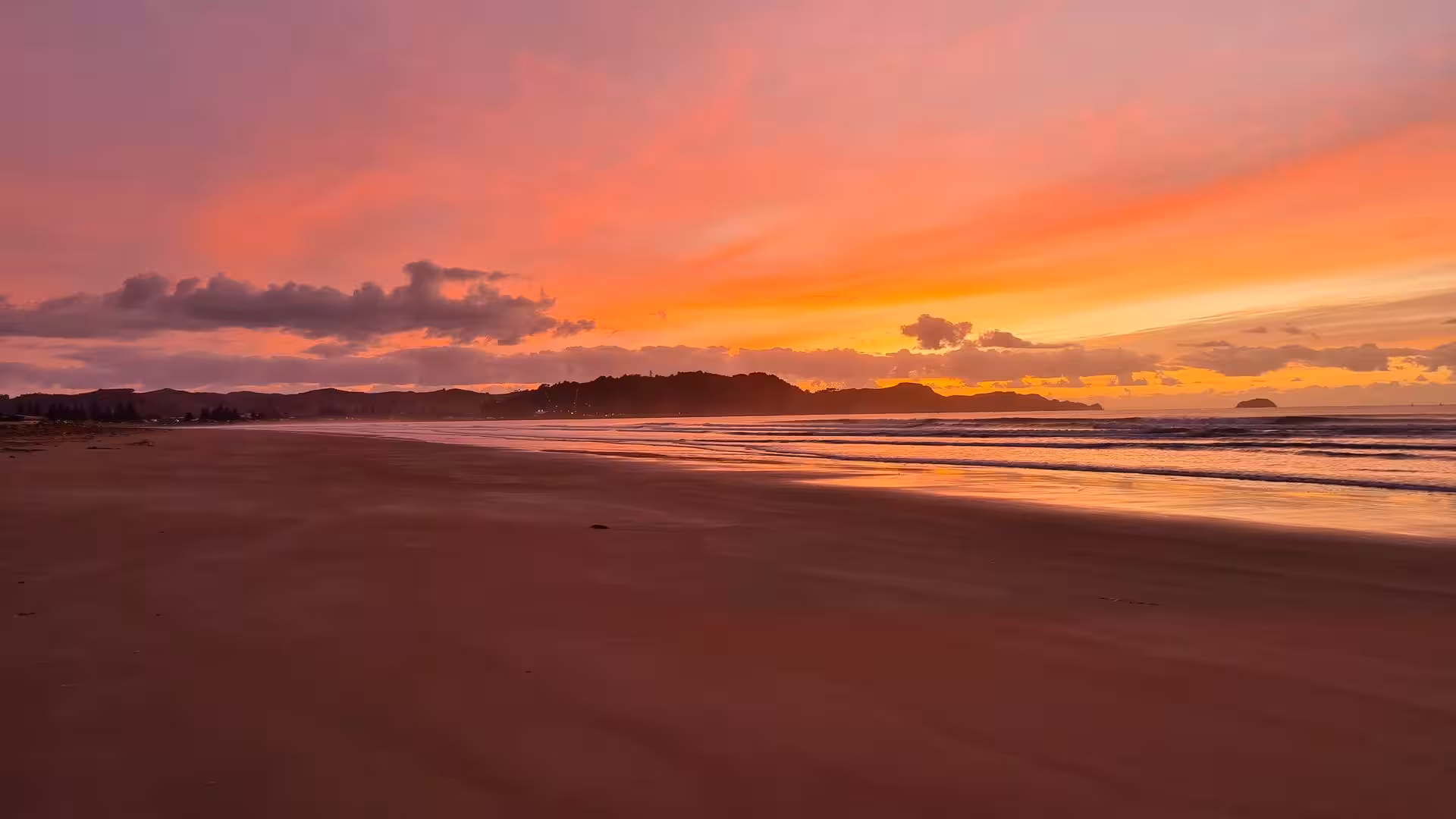 Orange and pink sky glows above waves on wet sand.