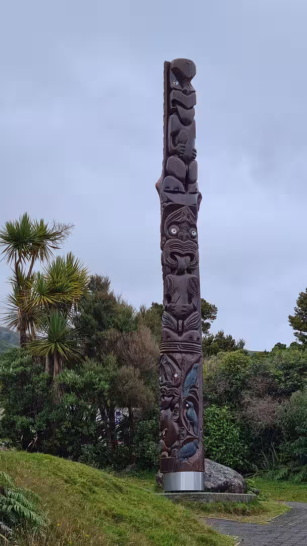 A tall carved native wooden pole with detailed figures rises from a lawn and garden.