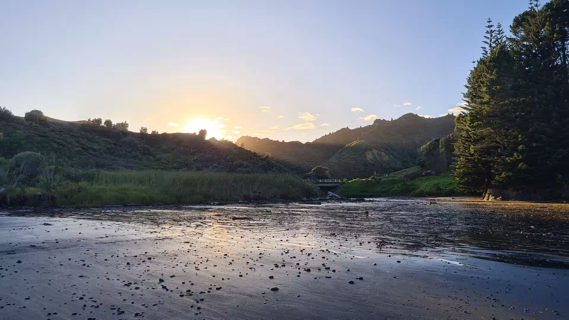 The sun dips between wooded hills above a glassy tidal river mouth.