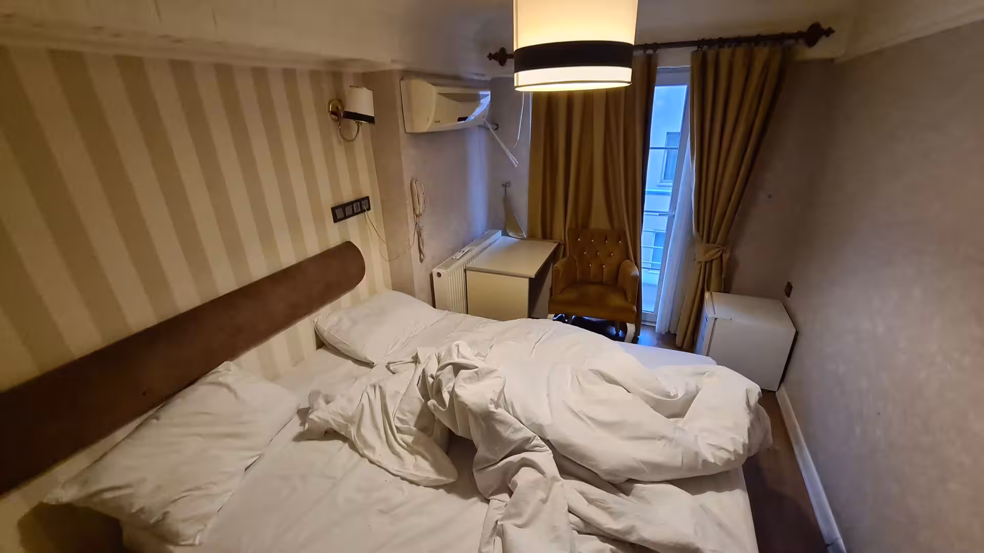 White sheets lie rumpled on a bed in a snug hotel room with striped walls.