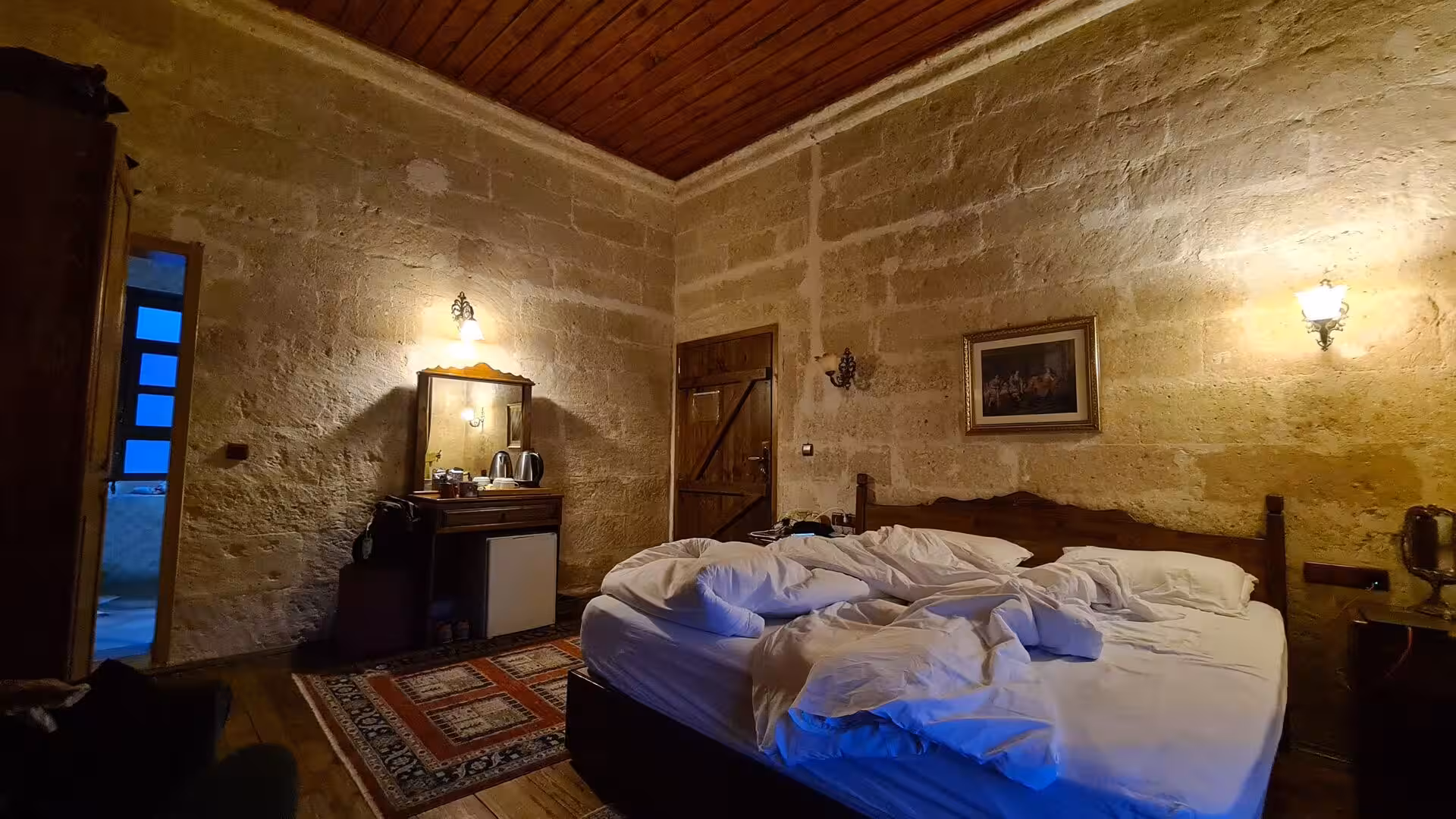 An unmade bed fills a warm stone-walled room.