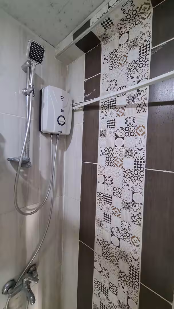 A white electric water heater sits beside a showerhead in a small tiled bathroom.