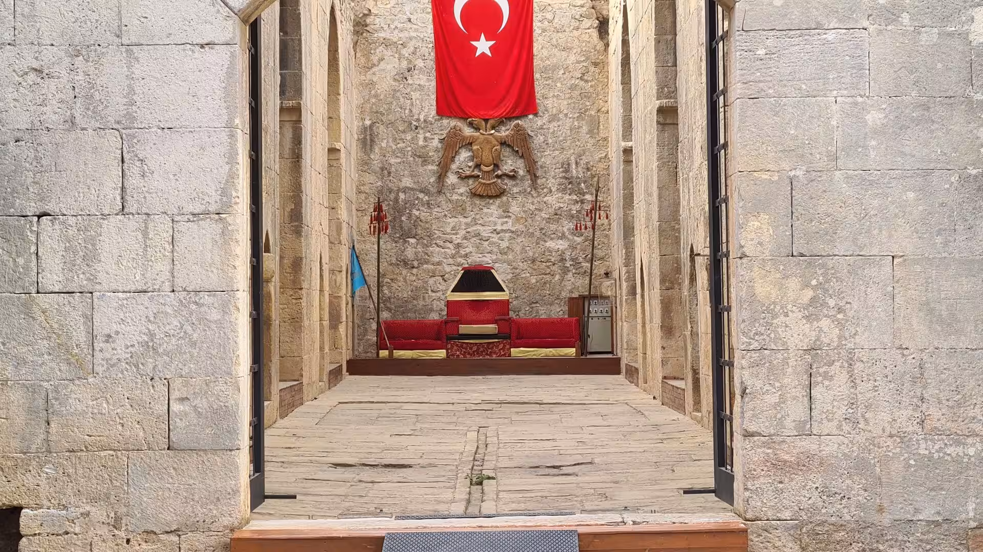 A crimson Turkish banner and a carved double-headed eagle watch over a red canopied dais in an old stone hall.