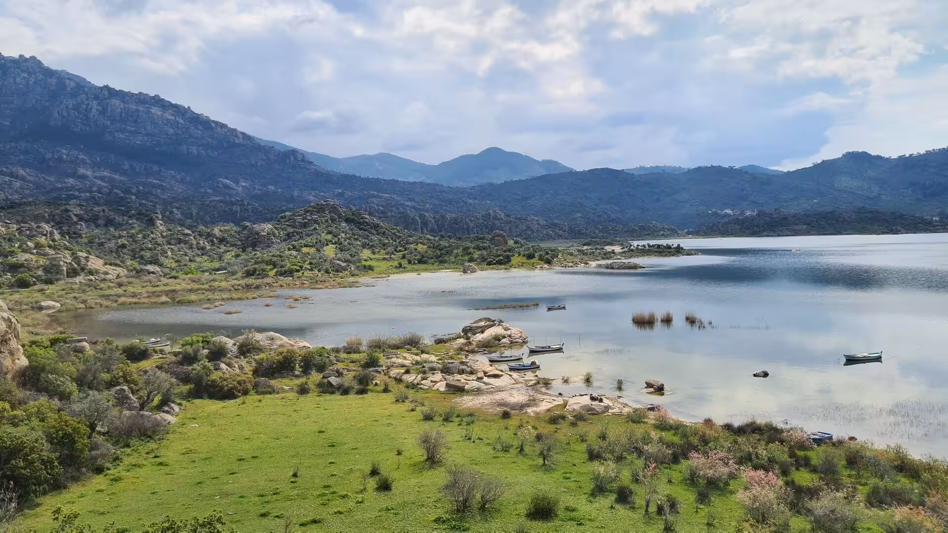 A quiet lake edged by green meadow and rugged hills, with small boats in the shallows.