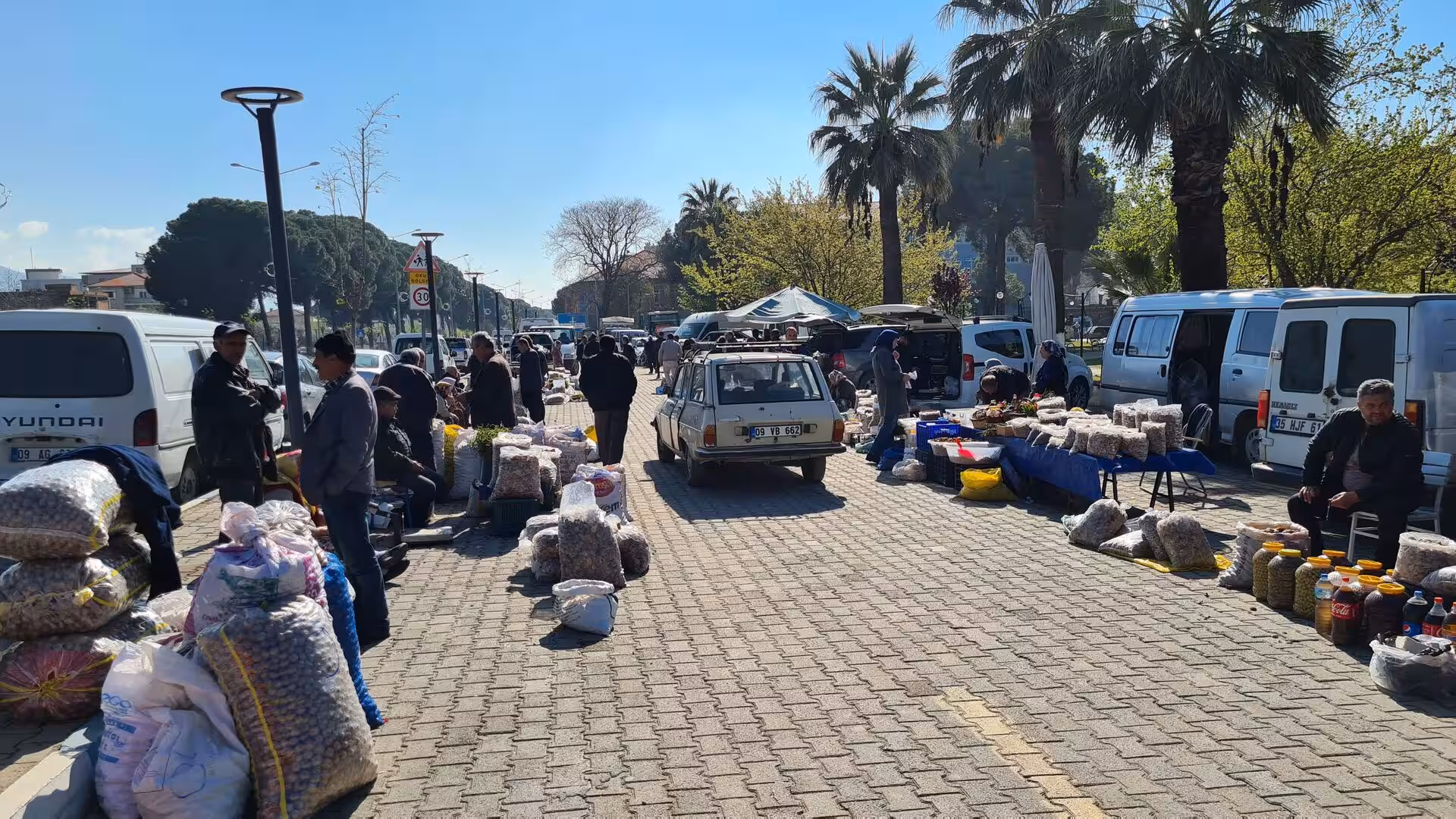 Vendors sell produce from sacks along a sunny market street.