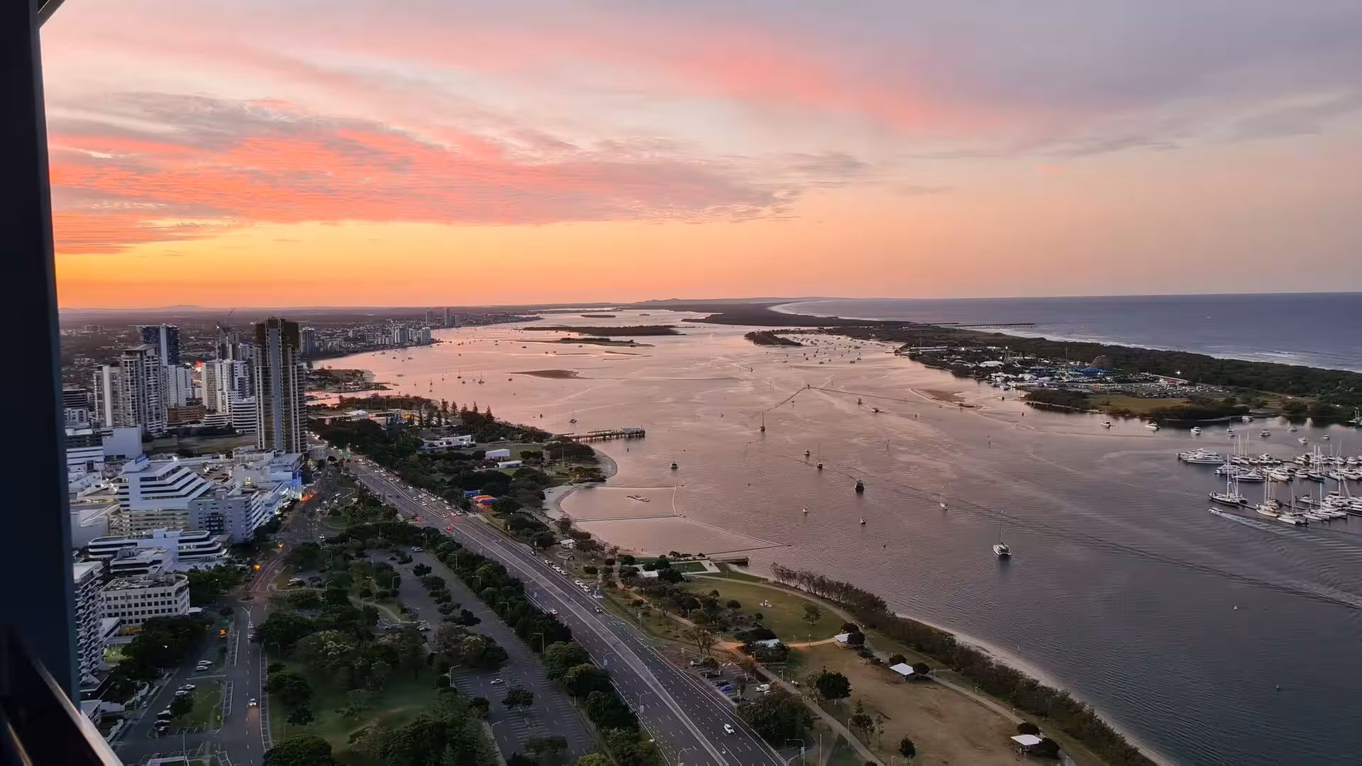 High-rise towers and a busy marina overlook an estuary glowing at sunset.