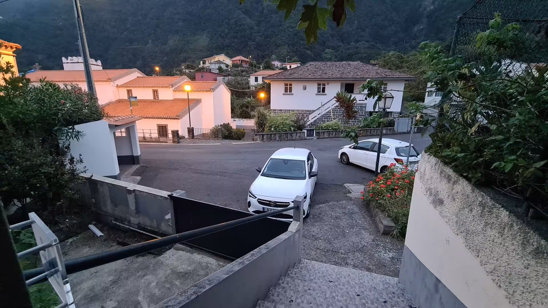 A quiet mountain lane with white houses, glowing lamps, and a lone car at twilight.