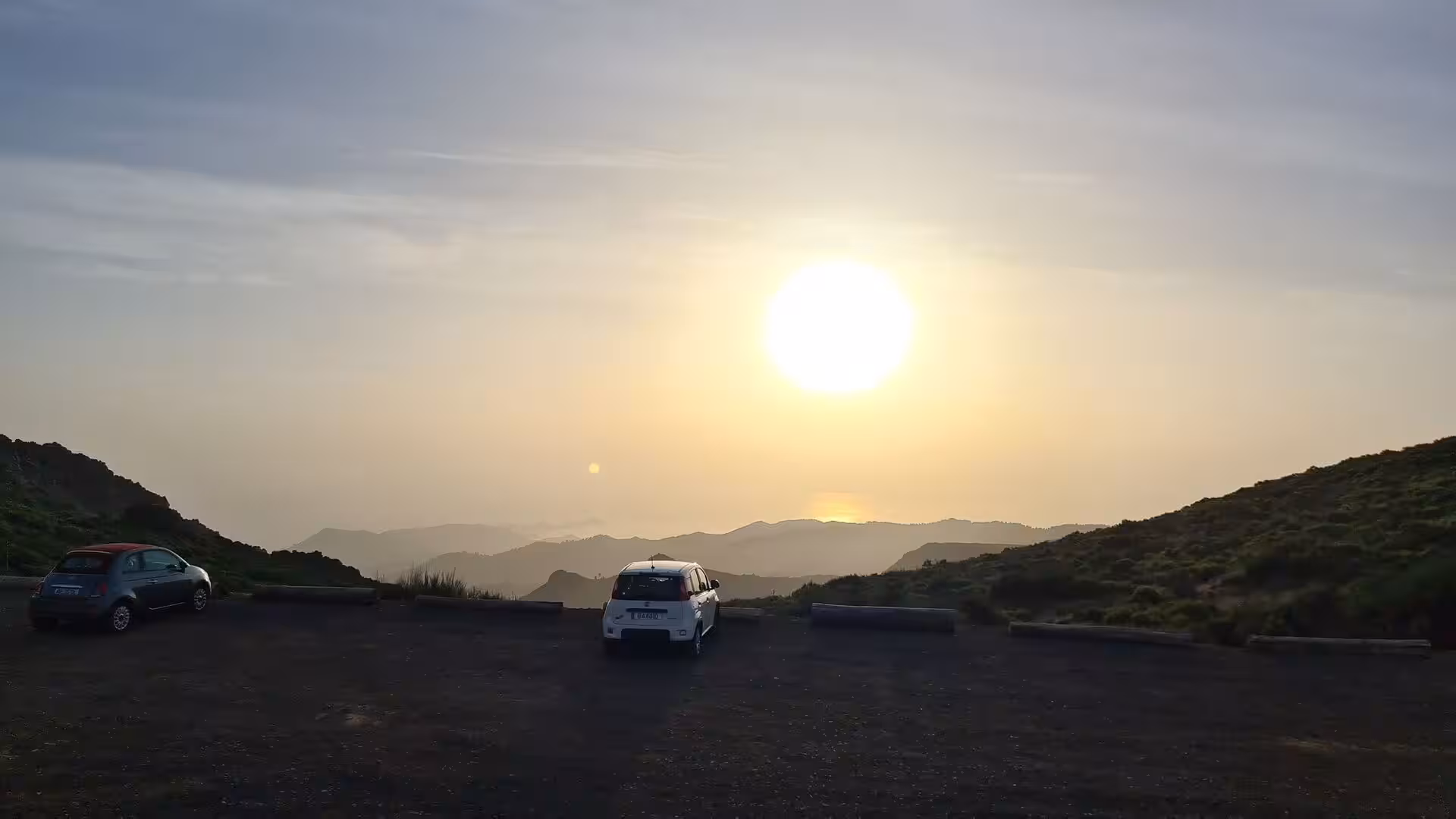 A white car and a dark hatchback sit on a high lookout beneath a huge setting sun.