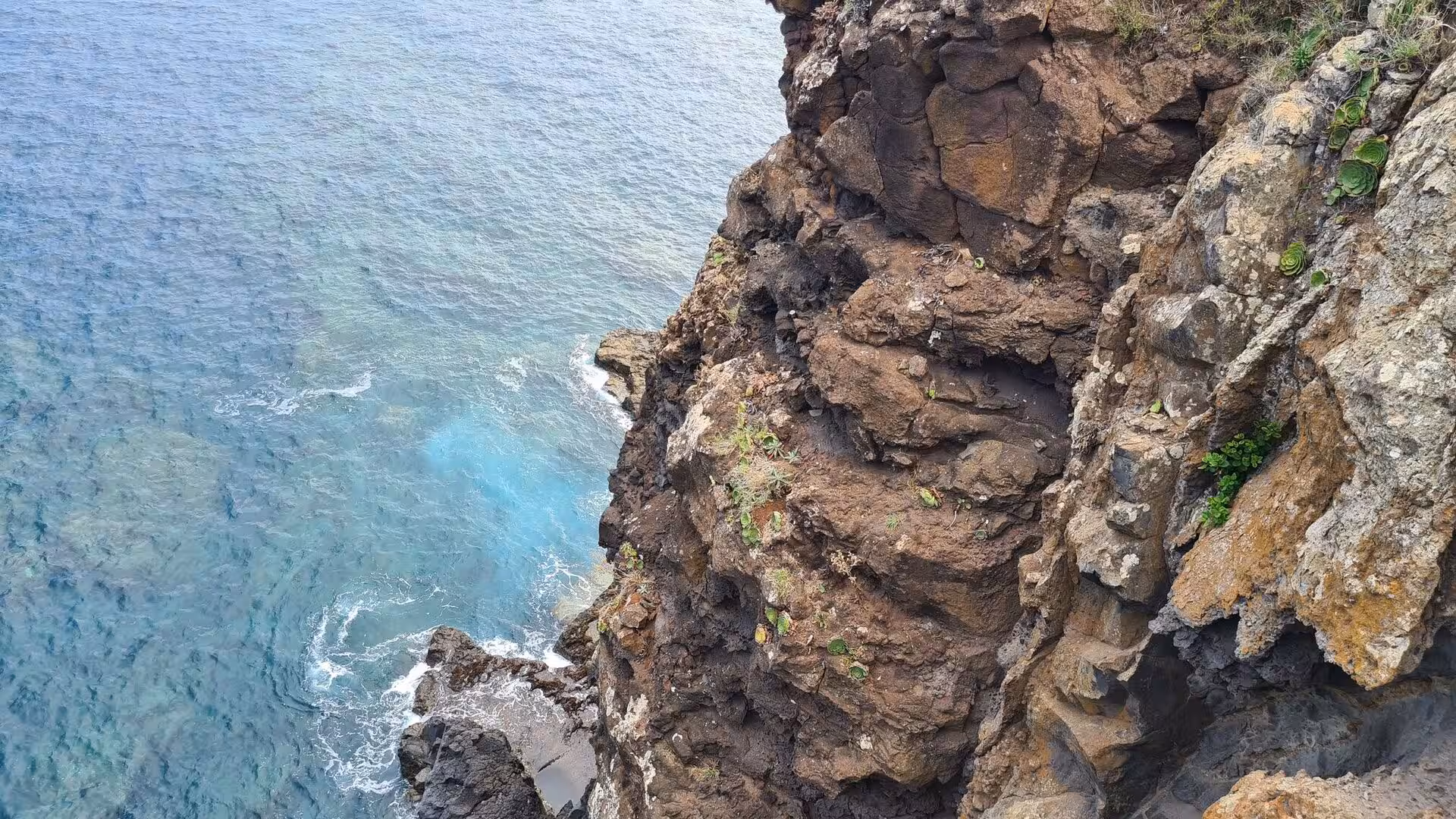 A steep weathered cliff drops straight into a teal sea.