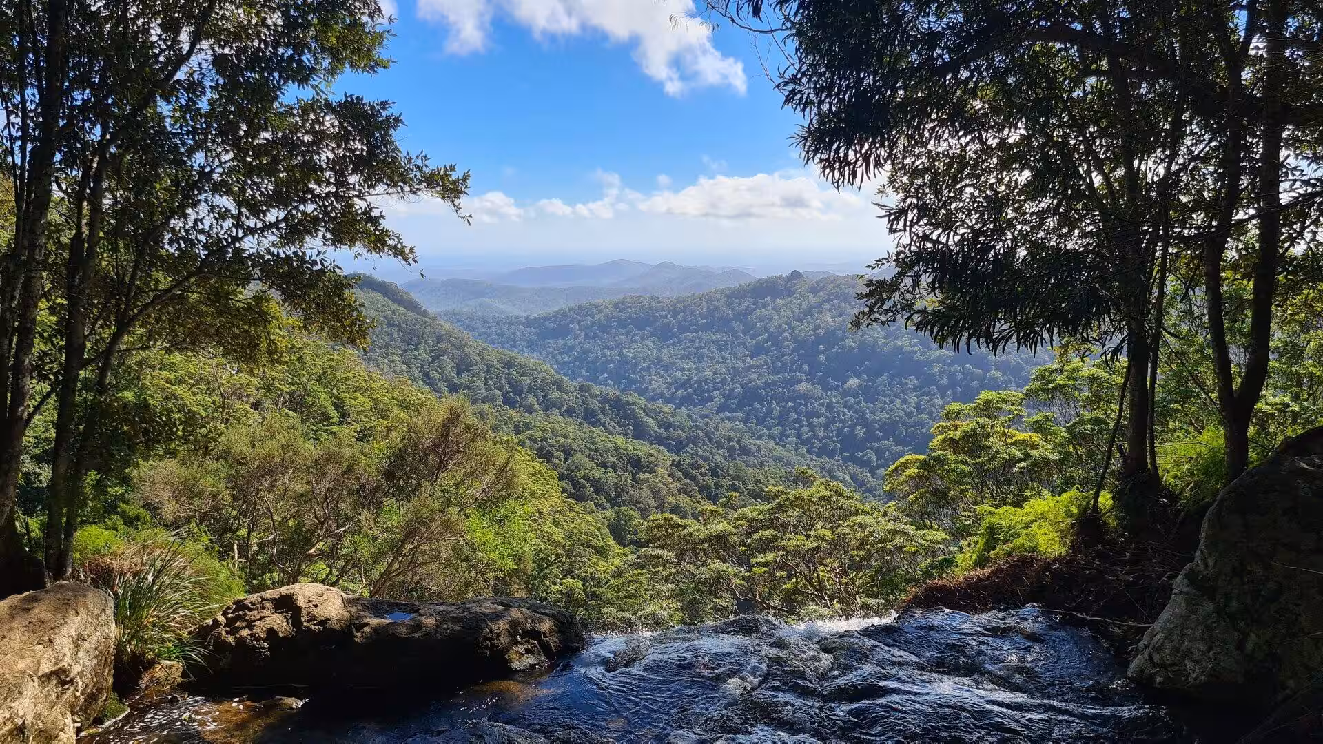 A stream flows through the foreground while forested hills roll toward distant mountains.